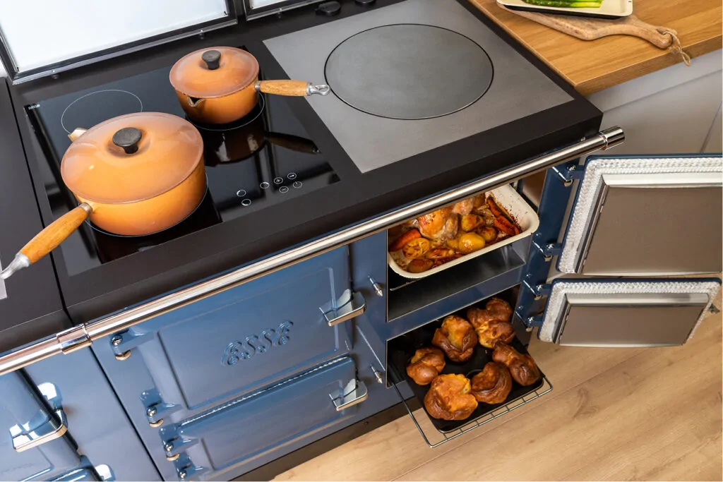 Een blauw ESSE elektrisch fornuis met een inductiekookplaat, waarop twee pannen staan en een geopende oven met geroosterde groenten en broodjes zichtbaar is.