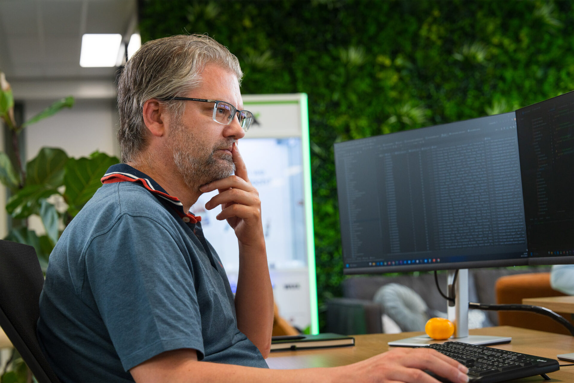 Wux-medewerker werkt gefocust met AI-data op twee schermen in een kantoor met groene plantenwand.
