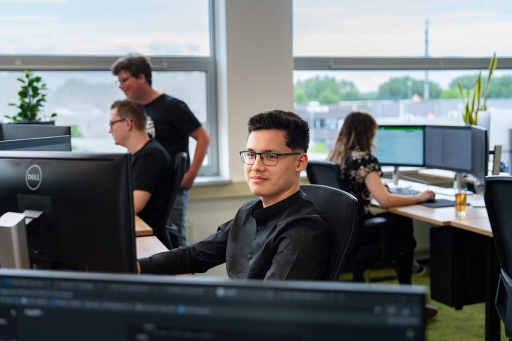 Een jonge man met bril en zwart overhemd zit ontspannen achter zijn computerscherm in een modern kantoor en kijkt met een tevreden blik. Op de achtergrond werken collega’s samen.