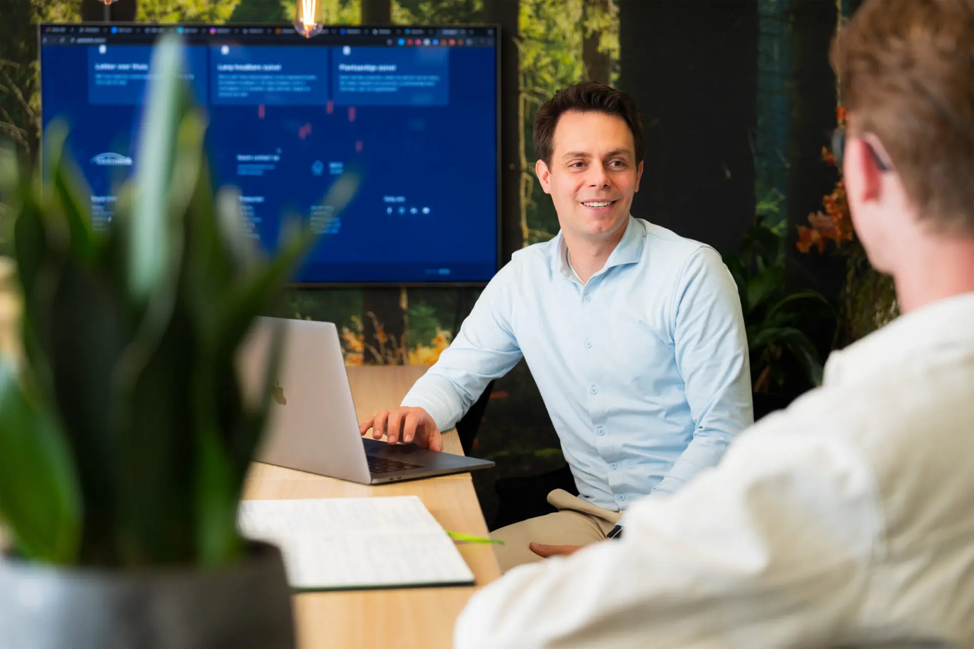 Man met bruine haren is een presentatie aan het geven aan zijn collega's met de presentatie op een TV scherm op de achtergrond