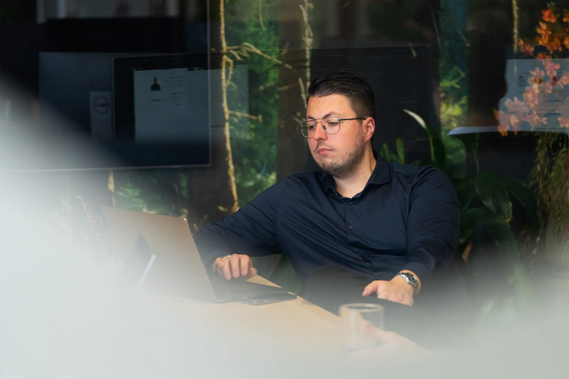 Man met bril werkt geconcentreerd op een laptop aan een bureau in een kantoorruimte met groene planten op de achtergrond.