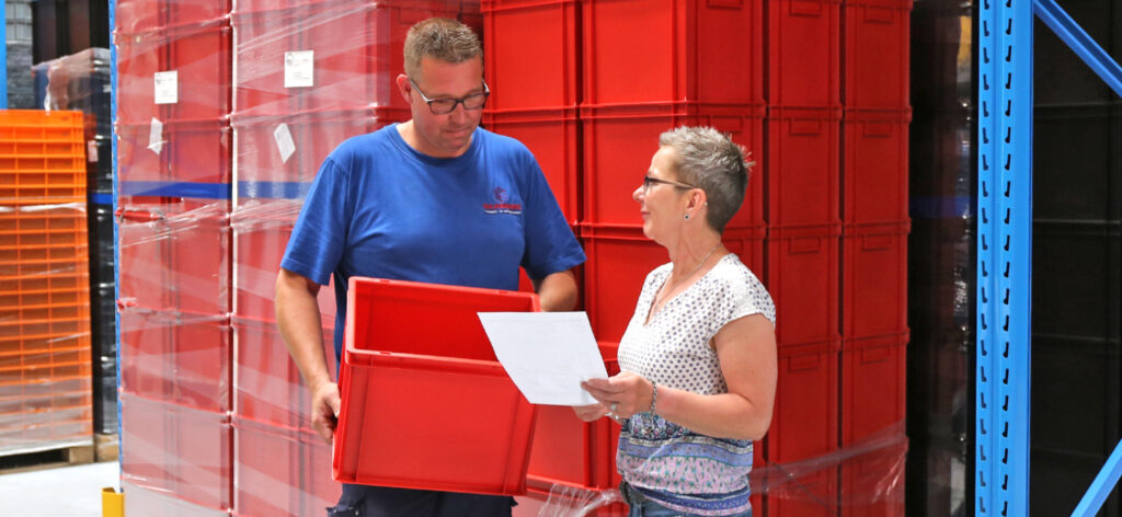 Magazijnmedewerkers bespreken logistieke details met rode kunststof bakken op de achtergrond.