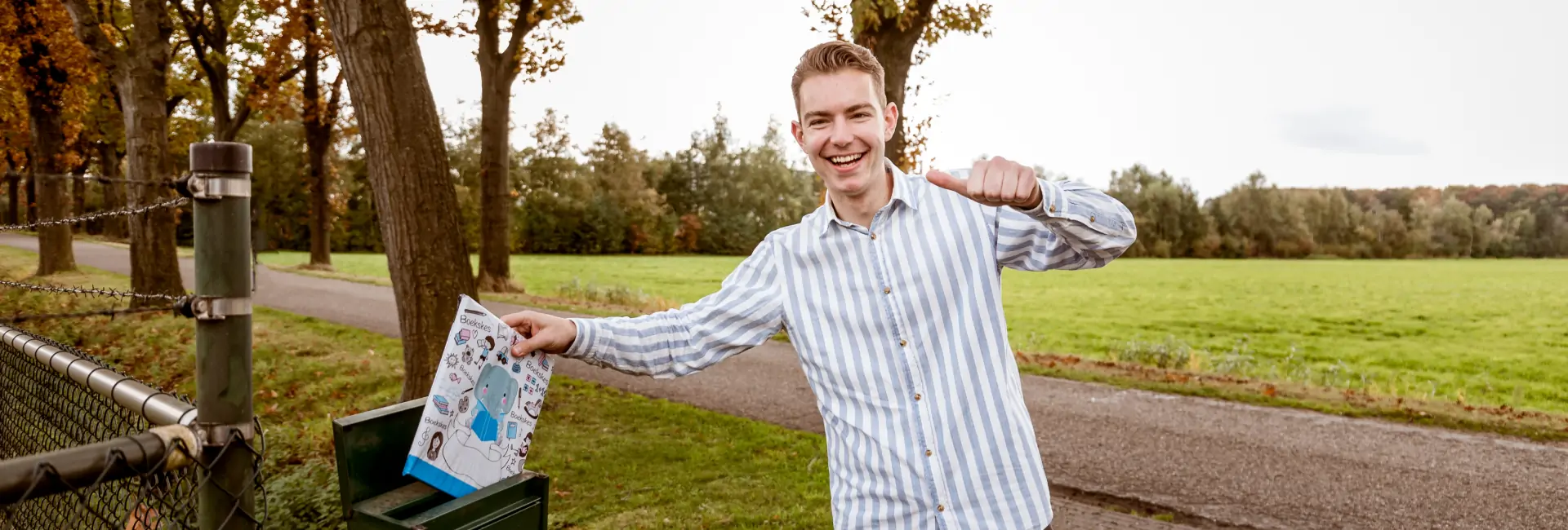 Een lachende jongeman stopt een Boekskes pakket in een brievenbus op het platteland, met een groene weide en bomen op de achtergrond.