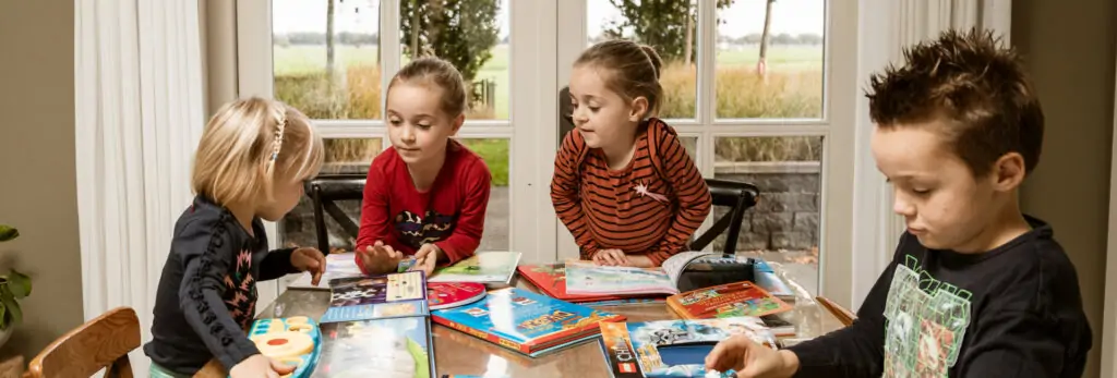 Vier jonge kinderen zitten samen aan een tafel en lezen verschillende kinderboeken, met een grote raamachtergrond die uitzicht biedt op een landelijke omgeving.
