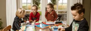 Vier jonge kinderen zitten samen aan een tafel en lezen verschillende kinderboeken, met een grote raamachtergrond die uitzicht biedt op een landelijke omgeving.