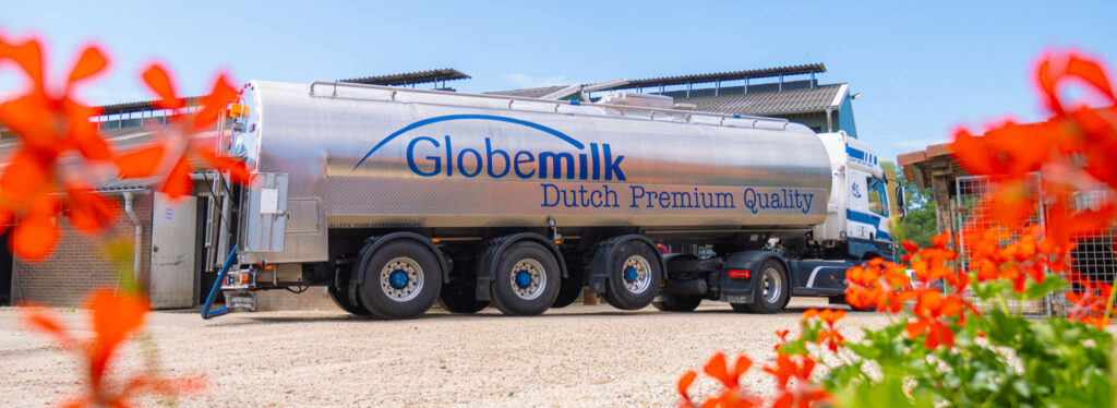 Een grote zilveren melktankwagen met het logo van Globemilk en de tekst 'Dutch Premium Quality', geparkeerd bij een boerderij, met rode bloemen op de voorgrond.