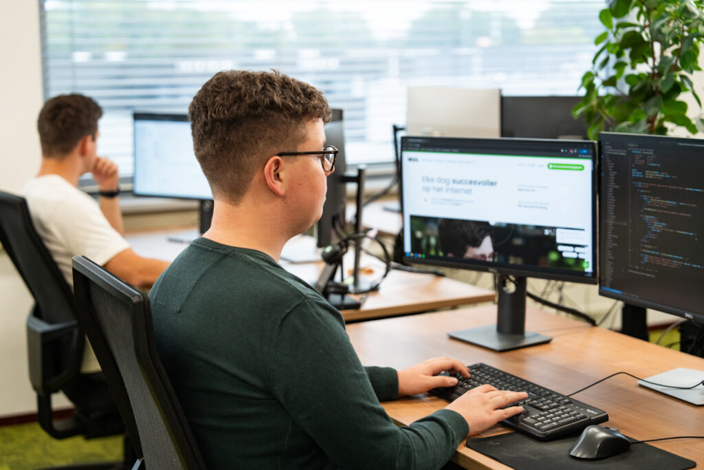 Ontwikkelaar werkt achter twee beeldschermen aan een website in het moderne kantoor van Wux.