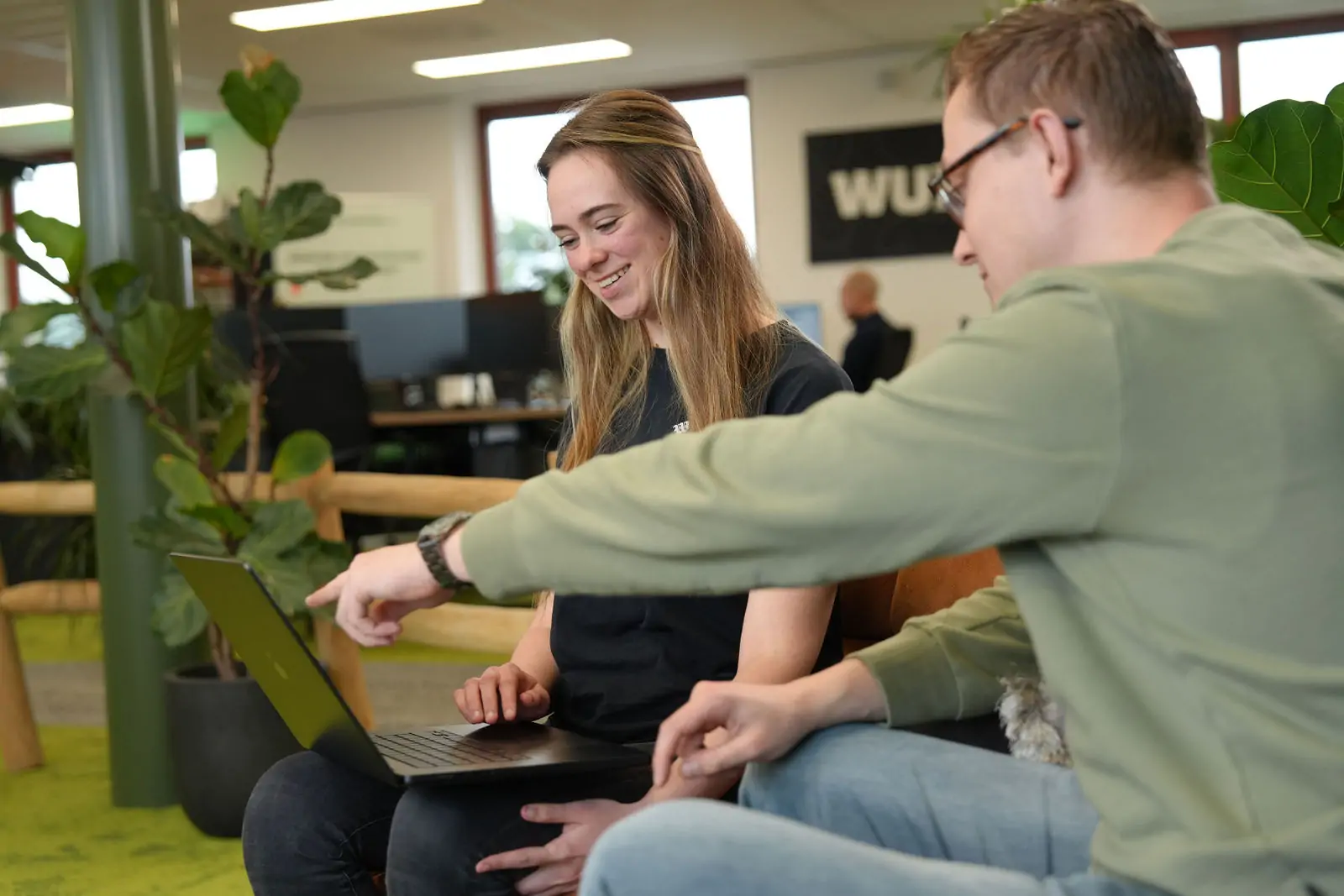 Twee collega’s in een modern kantoor, terwijl de man met een groene trui naar het laptopscherm wijst en de vrouw glimlachend meekijkt.