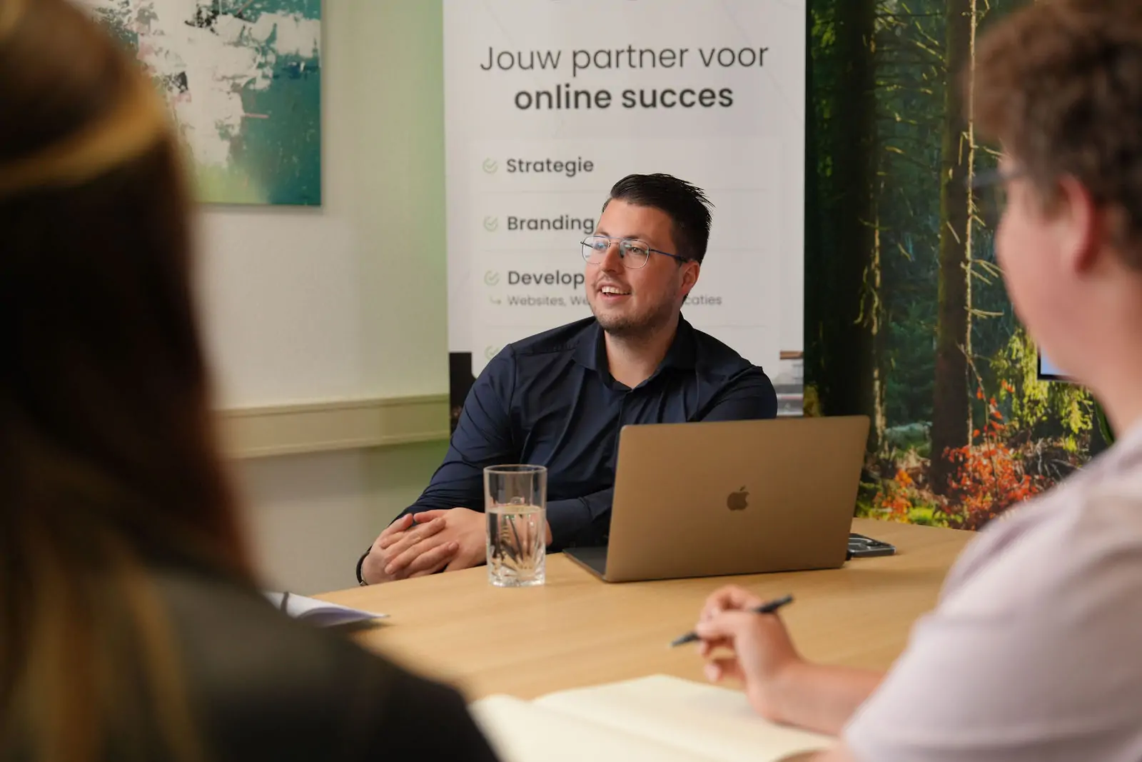 Man in donkerblauw overhemd zit achter zijn laptop in overleg met collega's