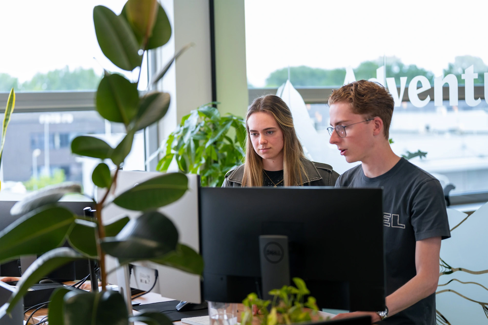 Twee collega’s werken samen achter een computerscherm in een modern en groen kantoor. De man, met bril en zwart T-shirt, legt iets uit terwijl de vrouw aandachtig luistert. Op de achtergrond zorgen grote ramen en planten voor een frisse en open werksfeer.