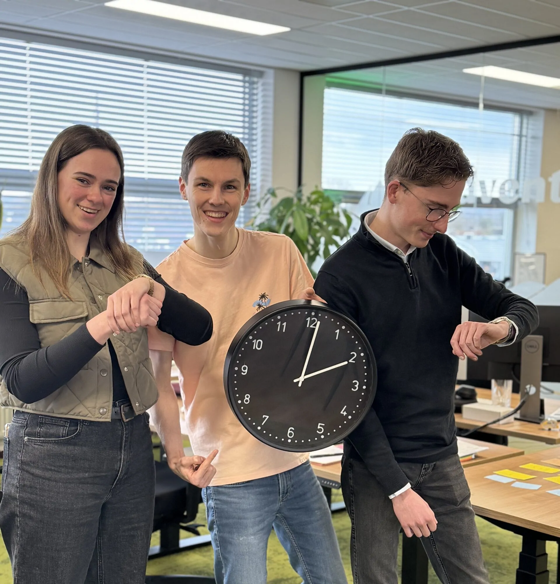 Drie collega’s in een kantoor kijken lachend naar de tijd, met een grote klok en horloges.