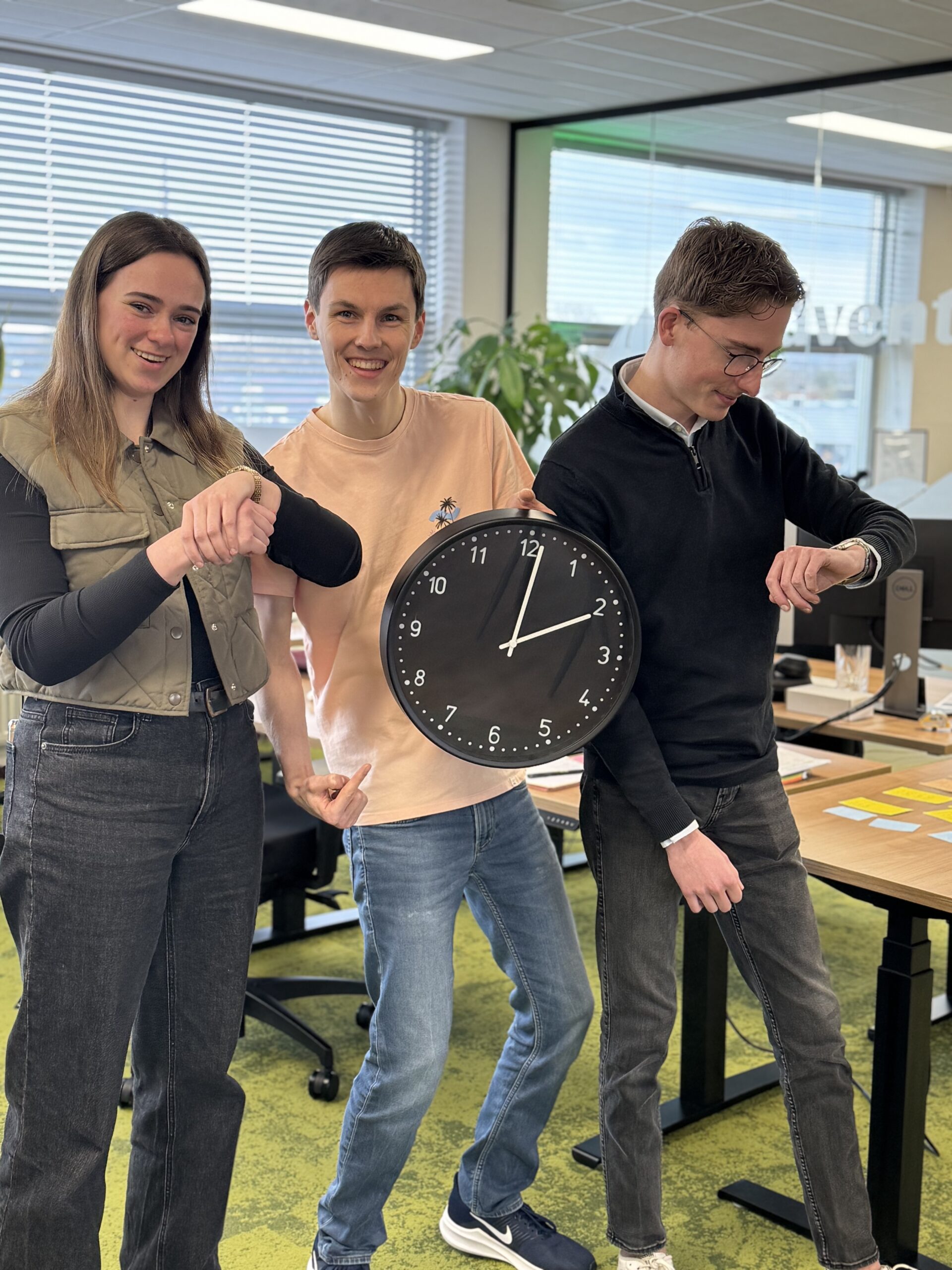Drie collega’s in een kantoor kijken lachend naar de tijd, met een grote klok en horloges.