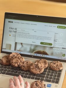 Een laptop met de website van Wux geopend waarop een cookie-melding te zien is. Op het toetsenbord liggen meerdere chocoladekoekjes