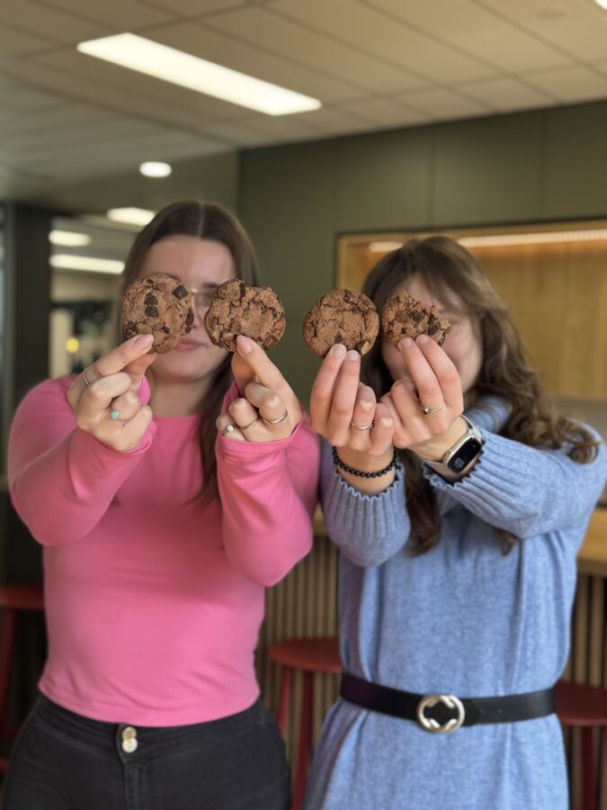 Twee jonge vrouwen houden vier chocoladekoekjes naar voren, waardoor hun gezichten grotendeels verborgen zijn. De vrouw links draagt een roze trui en de vrouw rechts een blauwe jurk met een zwarte riem. Ze staan in een modern ingerichte ruimte met groene en houten accenten