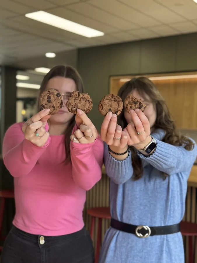 Twee jonge vrouwen houden vier chocoladekoekjes naar voren, waardoor hun gezichten grotendeels verborgen zijn. De vrouw links draagt een roze trui en de vrouw rechts een blauwe jurk met een zwarte riem. Ze staan in een modern ingerichte ruimte met groene en houten accenten