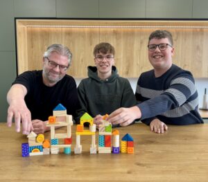 Drie mannen zitten samen aan een houten tafel en bouwen lachend een toren van kleurrijke houten blokken.