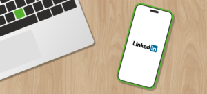 Een smartphone met het LinkedIn-logo op het scherm ligt schuin op een houten tafel, naast een laptop met zwarte en groene toetsen