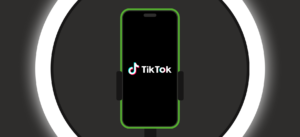 Een smartphone met een groen randje is bevestigd in een houder voor een ringlamp en toont het TikTok-logo, wat een setting suggereert voor het maken van content.