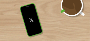 Een smartphone met een groen randje ligt op een houten tafel en toont het logo van X (voorheen Twitter) op het scherm. Ernaast staat een kop koffie
