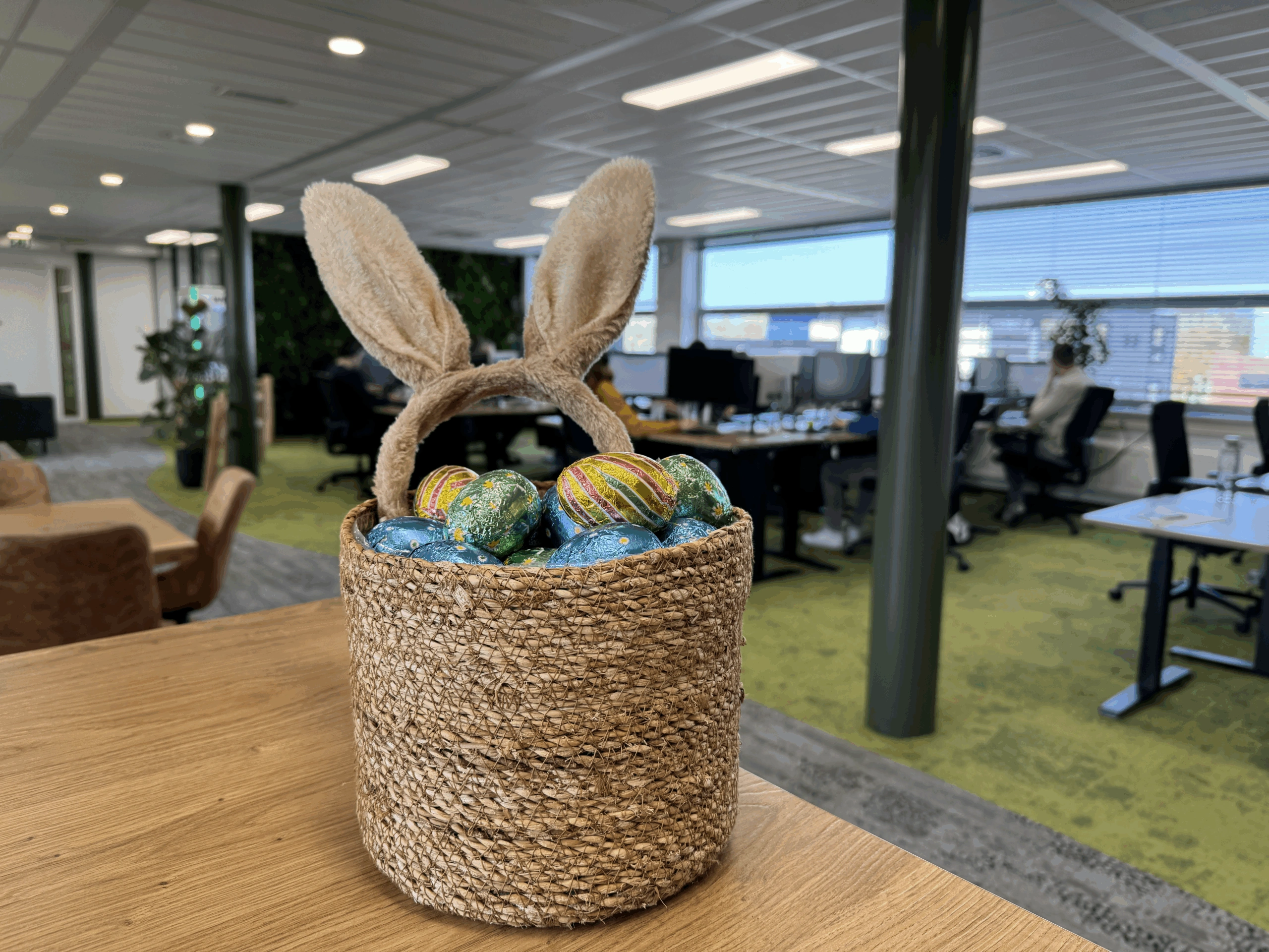 Een rieten mandje met paaseitjes en konijnenoren staat op een houten tafel in het moderne kantoor van Wux.