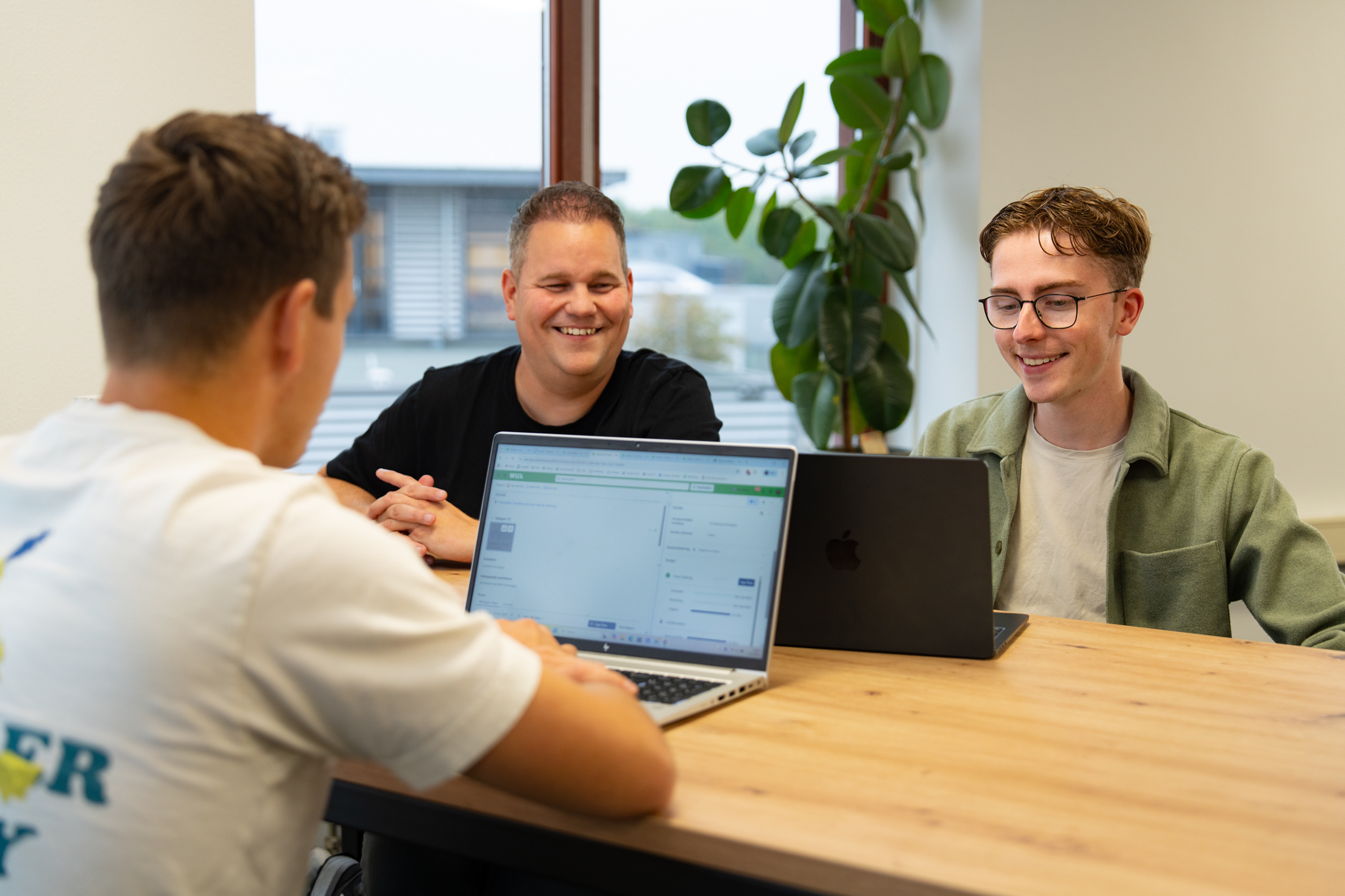 Drie collega’s overleggen met laptops aan configurators aan tafel in een lichte kantoorruimte met planten