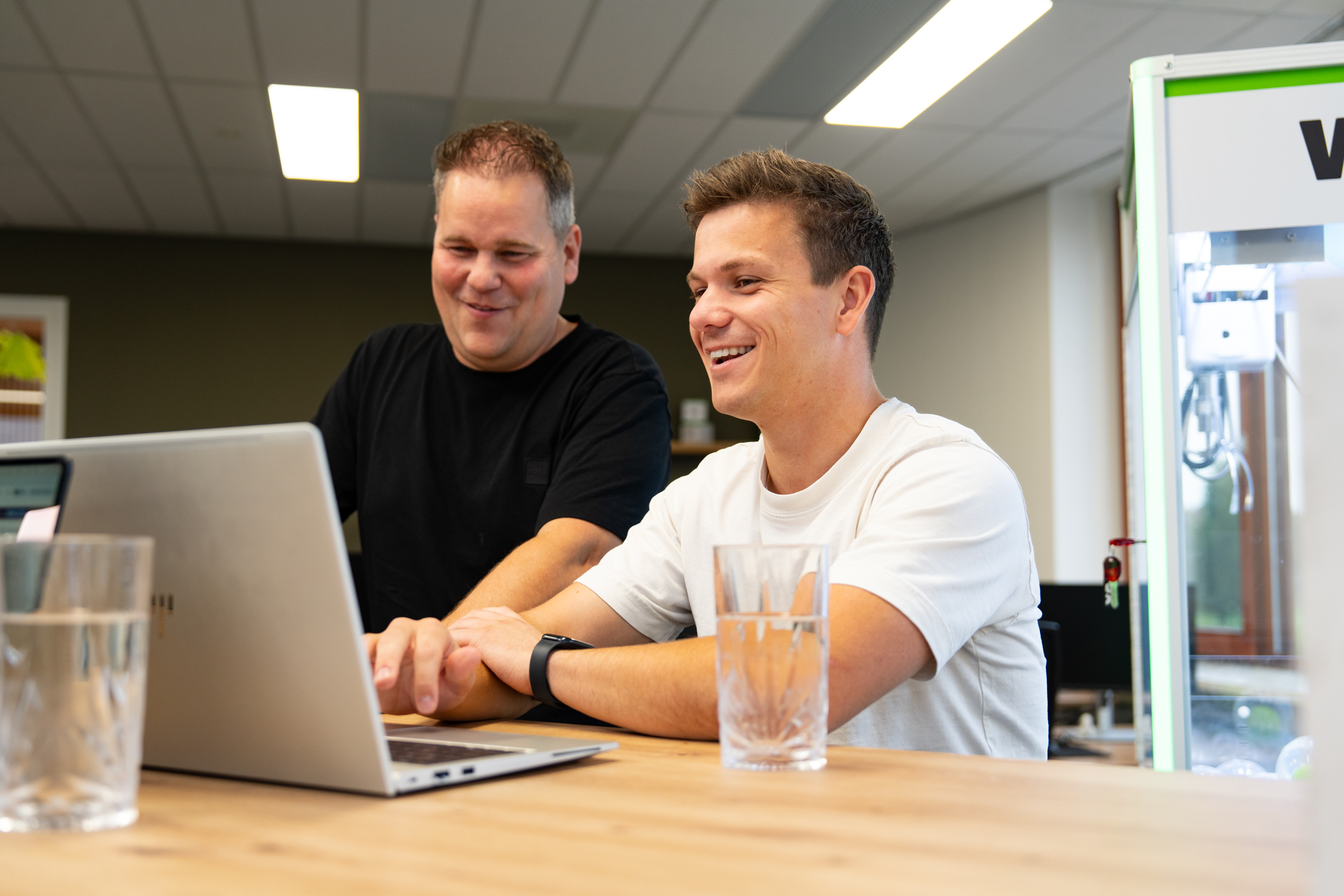 Twee collega’s werken samen achter een laptop en lachen tijdens een overleg in het moderne kantoor van Wux.