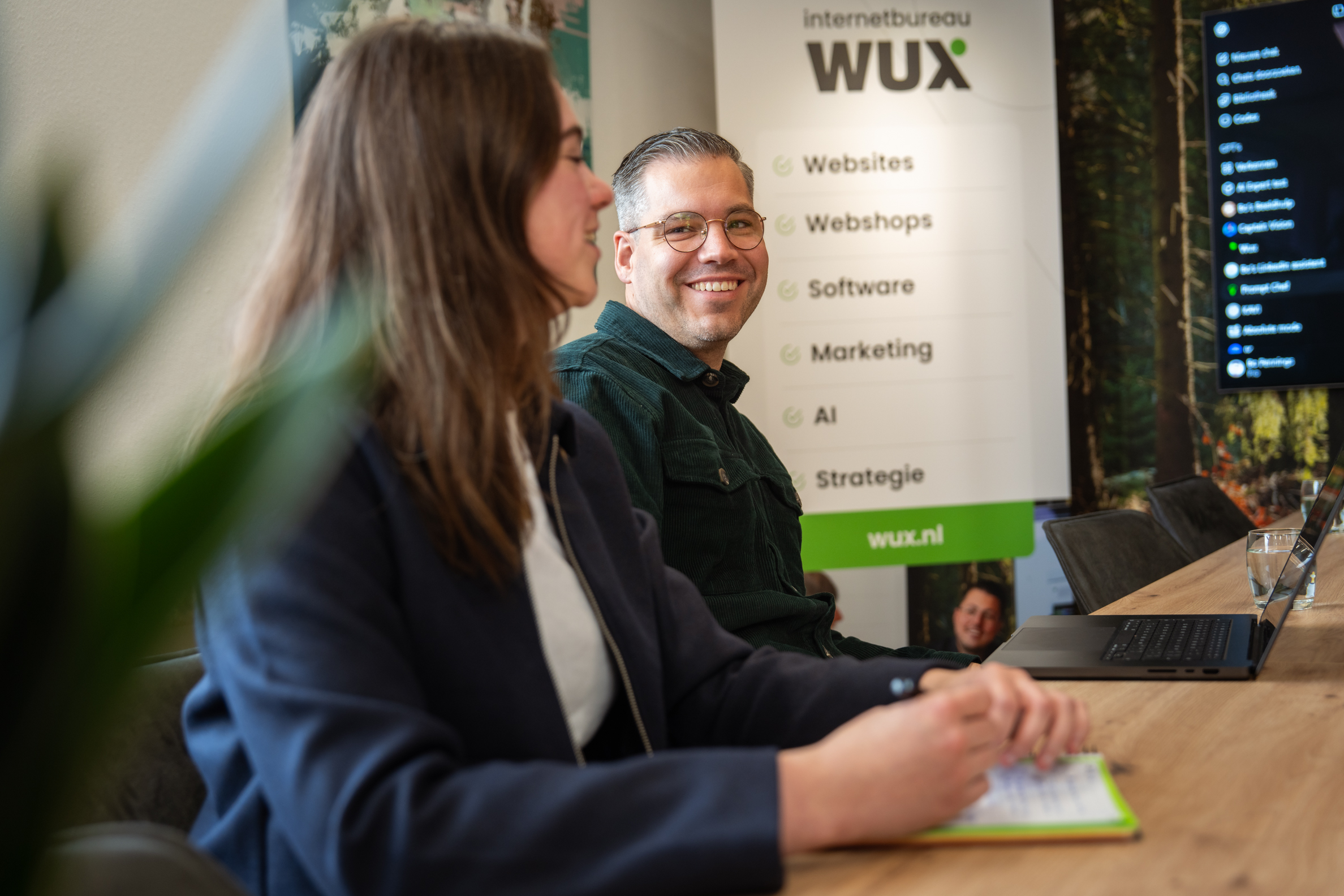 Twee collega’s tijdens een overleg aan tafel bij internetbureau WUX, met branding zichtbaar op de achtergrond.
