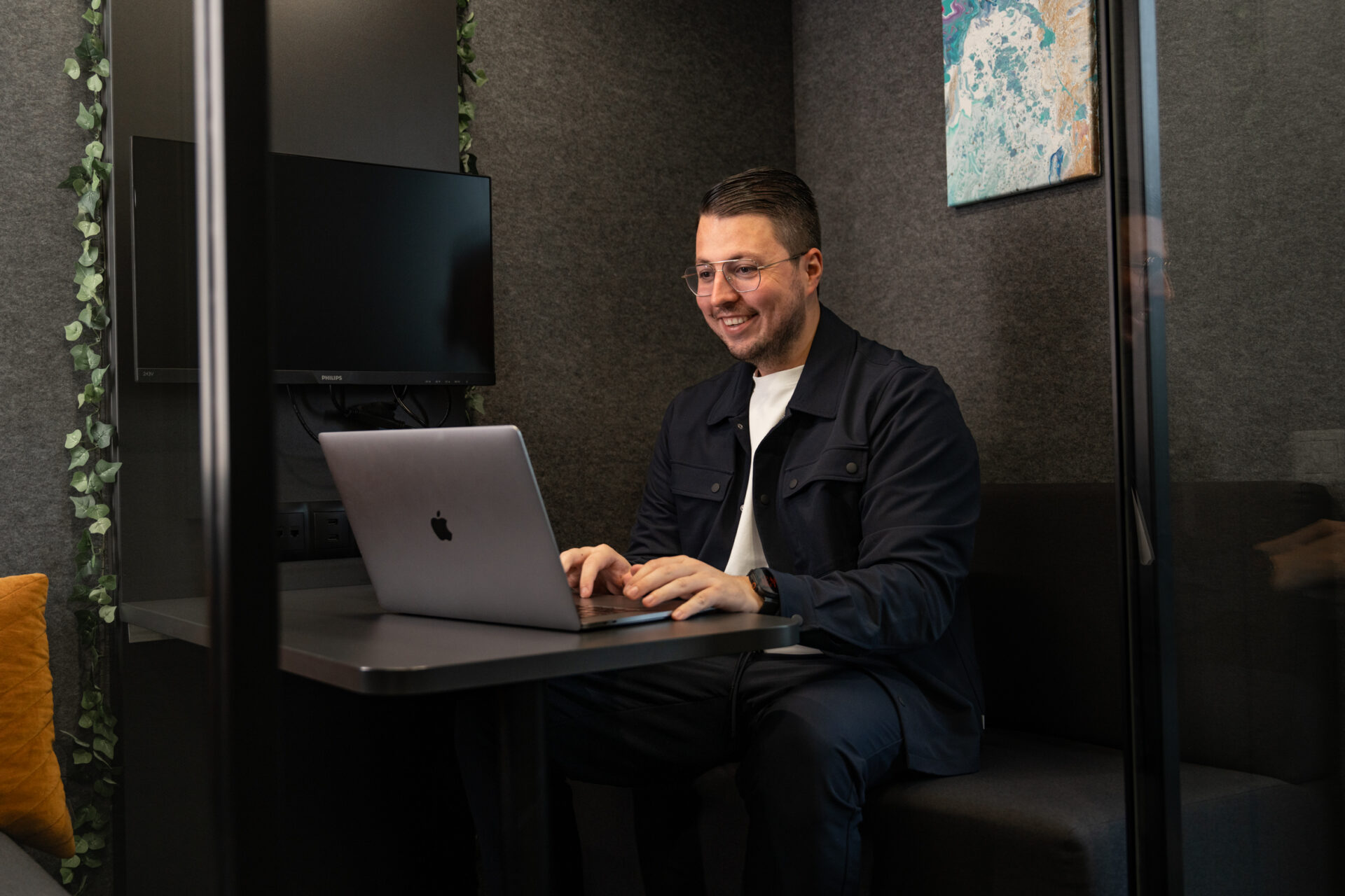 Man werkt glimlachend op zijn laptop in een stille werkruimte met moderne inrichting