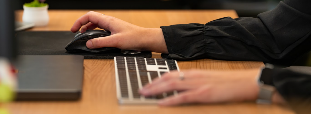 Close-up van een medewerker die achter een houten bureau met muis en toetsenbord op een computer werkt.