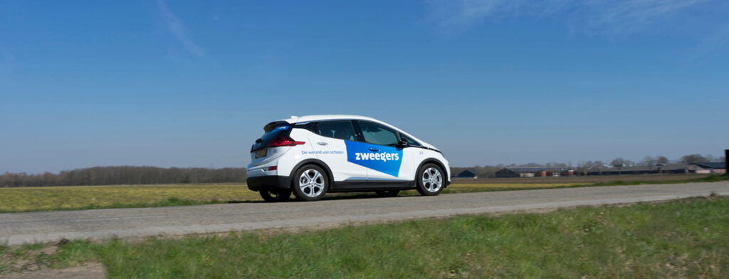 Onderweg naar de volgende klus: een witte bedrijfsauto met het blauwe Zweegers-logo en de tekst 'De wereld van schoon' rijdt over een landelijke weg onder een strakblauwe hemel.
