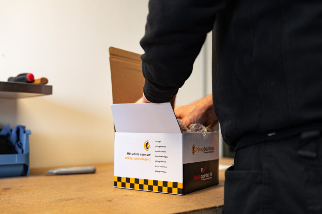 Een medewerker in donkere werkkleding plaatst een in noppenfolie verpakt onderdeel in een bedrukte verzenddoos van Airbagbank.eu op een werkbank.