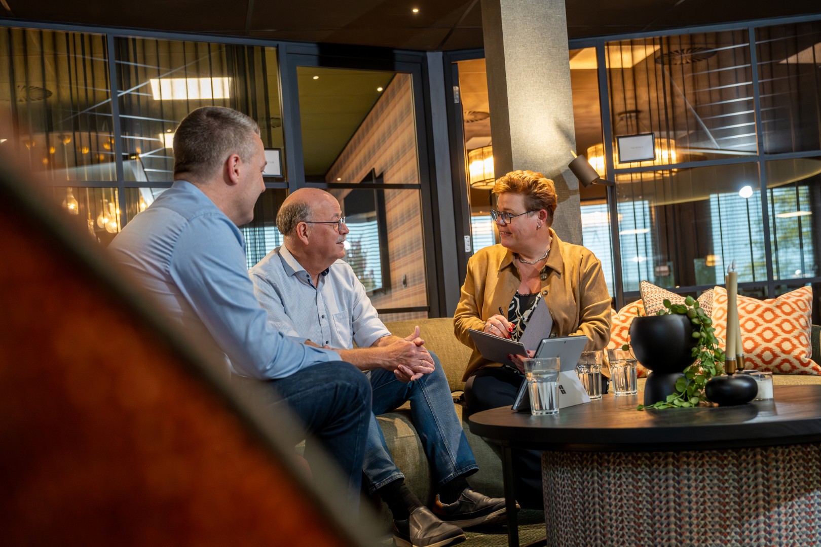 Persoonlijk ICT-adviesgesprek bij ADA ICT: een adviseur met een tablet in overleg met twee personen in een sfeervolle, informele setting.
