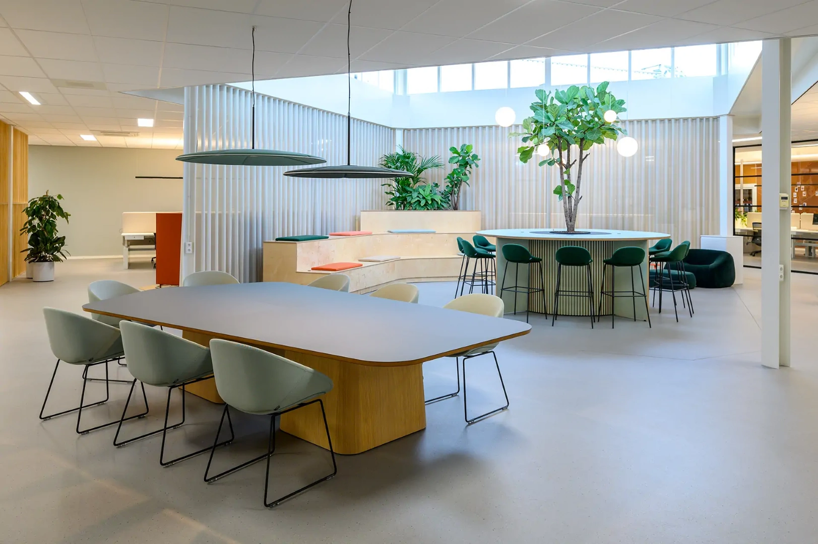 Een lichte, open kantoorruimte ingericht door Lande. Op de voorgrond staat een ruime vergadertafel met lichtgroene kuipstoelen. Op de achtergrond bevindt zich een houten zittribune met gekleurde kussens en een hoge, ronde ontmoetingstafel met een grote boom in het midden, omringd door donkergroene barkrukken.