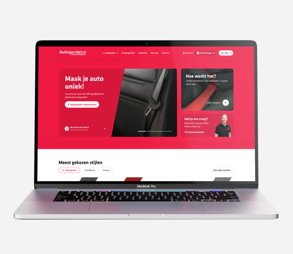 Een frontaal aanzicht van een MacBook Pro laptop die de rode homepage van Autogordel.nl toont. Het scherm bevat de tekst "Maak je auto uniek!" met een knop "Autogordels samenstellen". Daarnaast zijn er secties voor reviews, uitleg-video's, contactgegevens en "Meest gekozen stijlen" van gordelbanden.