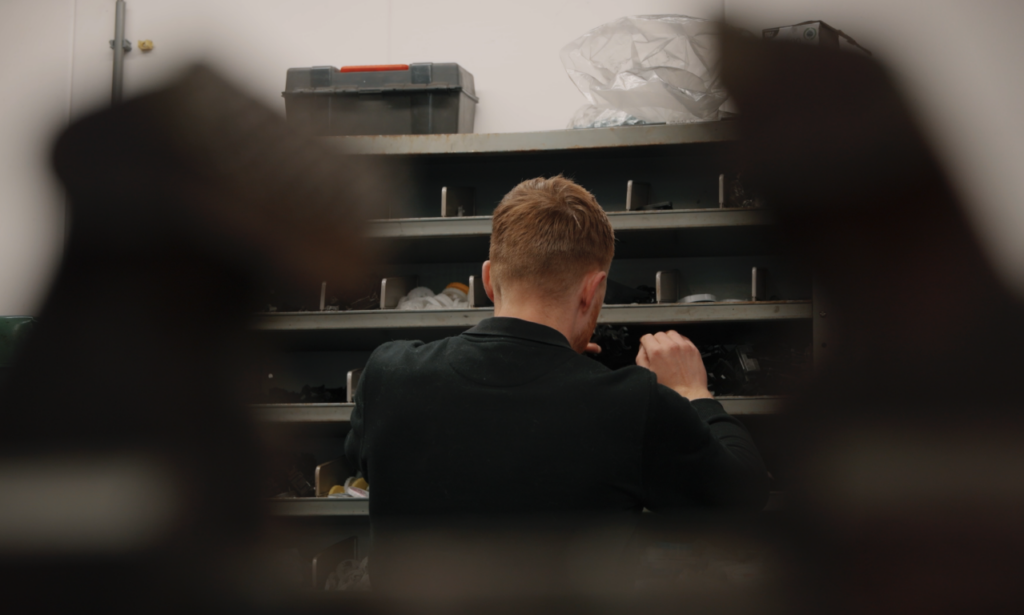 Op de rug gezien staat een medewerker in donkere werkkleding auto-onderdelen te zoeken of te pakken uit een grijze vakkenkast.