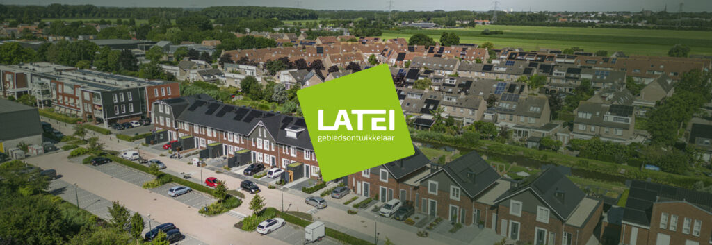 Brede, panoramische luchtfoto van een door LATEI ontwikkelde woonwijk. De wijk bestaat uit diverse moderne eengezinswoningen, merendeels voorzien van zonnepanelen, gelegen aan een overzichtelijke straat met parkeervakken en groen. Exact in het midden van de foto is een groot groen vlak geplaatst met daarin het witte logo van 'LATEI gebiedsontwikkelaar'.