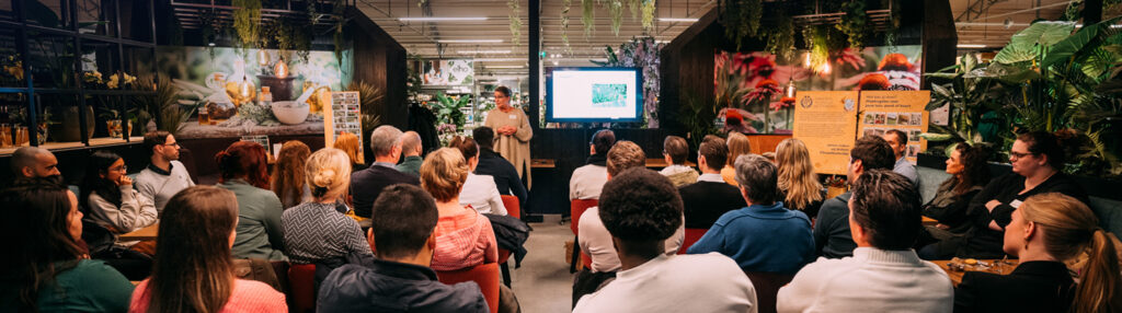 Een foto van een workshop of presentatie in een intieme, met planten gevulde showroom of binnentuin. Een vrouwelijke spreker staat voorin en presenteert een dia op een groot scherm aan een publiek van ongeveer dertig mensen die luisteren en kijken. De ruimte heeft een warme, groene sfeer met hangplanten en grote botanische wandafdrukken.